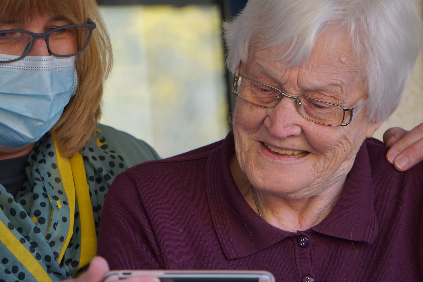 Une femme âgée manipulant un smartphone avec l'aide d'une infirmière portant un masque de protection.
